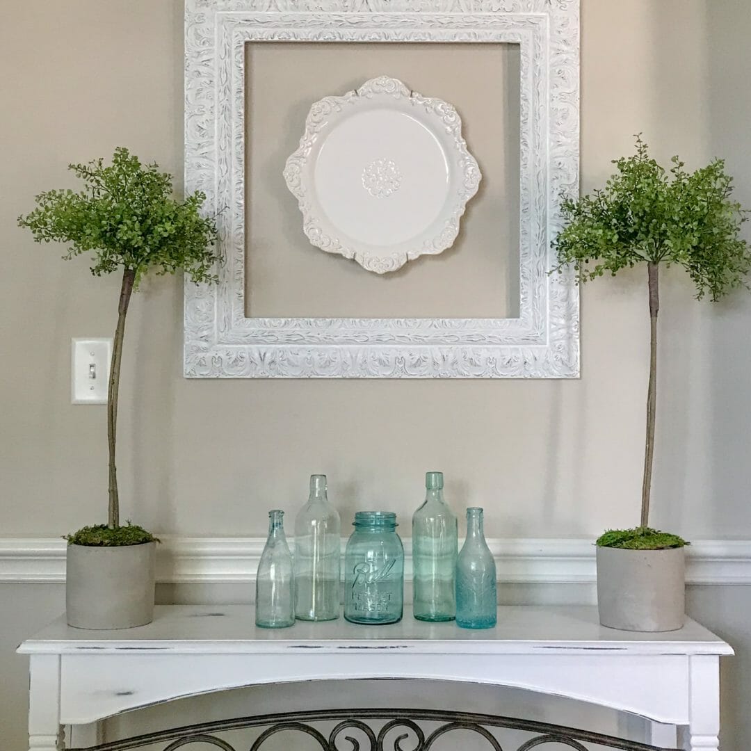 Console table with topiaries, blue bottles and white charger plate inside white frame