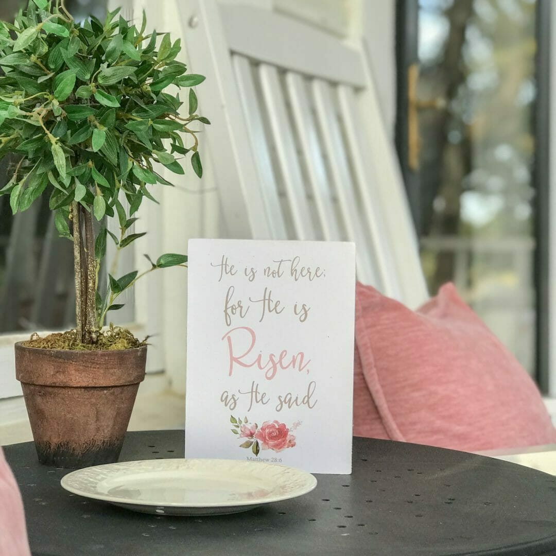 He is Risen sign sitting on table with topiary and white chair with pink pillow in background