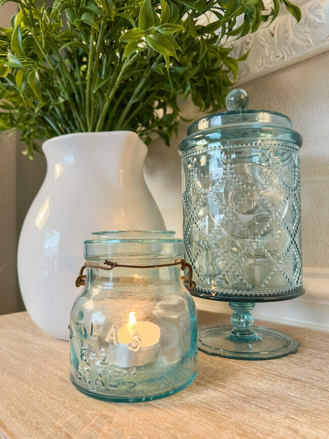 white pitcher with green stems and two blue jars. one with a candle