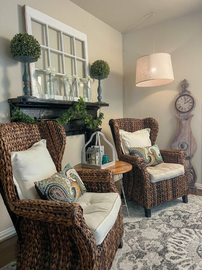 sitting are with rattan chairs, standing clock and black mantel with white window pane and flowers