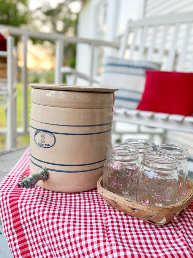 Vintage crock with spigot and 4 glass mason jars on top of red and white checked napkin