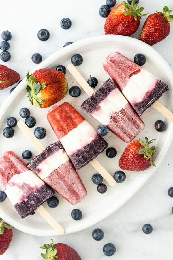 red, white and blue popsicles with blueberries and strawberries on white platter