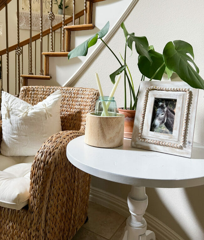 white table, rattan chair with white pillow and plant with frame on table