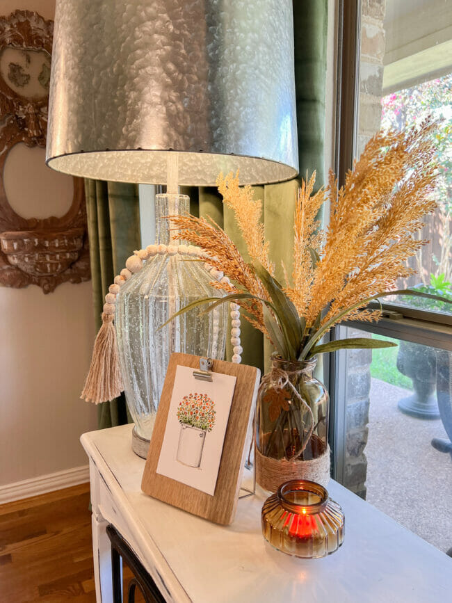 Table with lamp, wheat and watercolor print clipped to frame