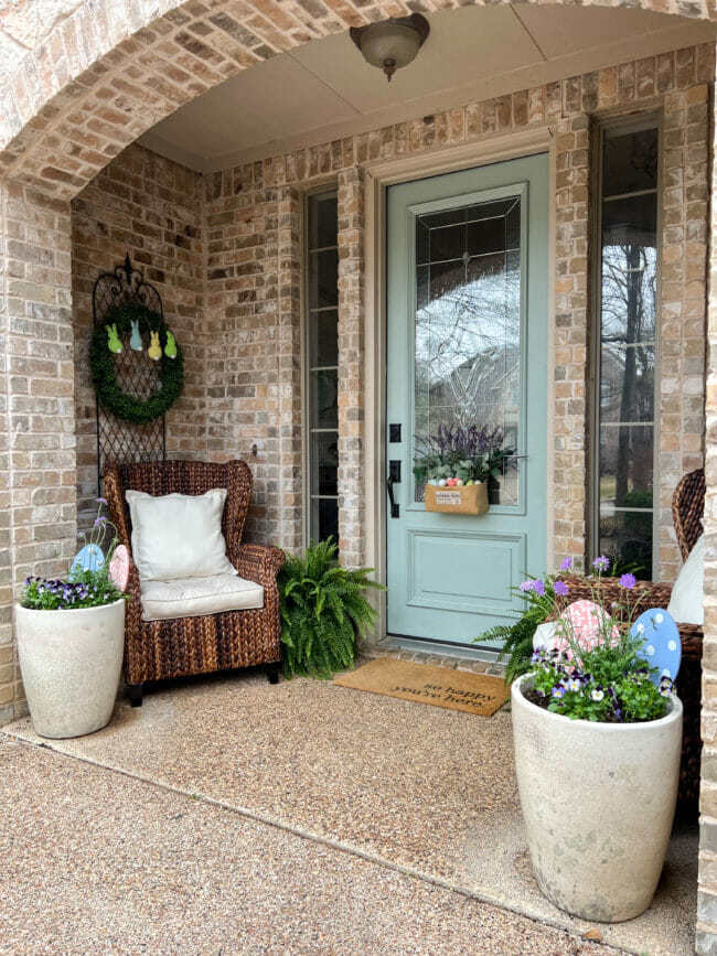 pale blue/green door with burlap basket of flowers, white pots with flowers and eggs and brown rattan chairs