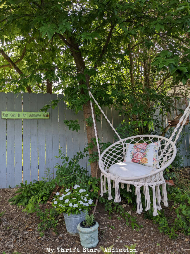 macrame swing hanging in tree with potted flowers and gray fence