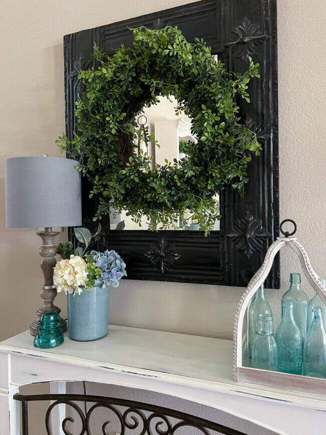 boxwood wreath hanging on black mirror with blue bottles, lamp and flowers