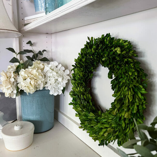 boxwood wreath hanging in hutch with blue crock and hydrangeas