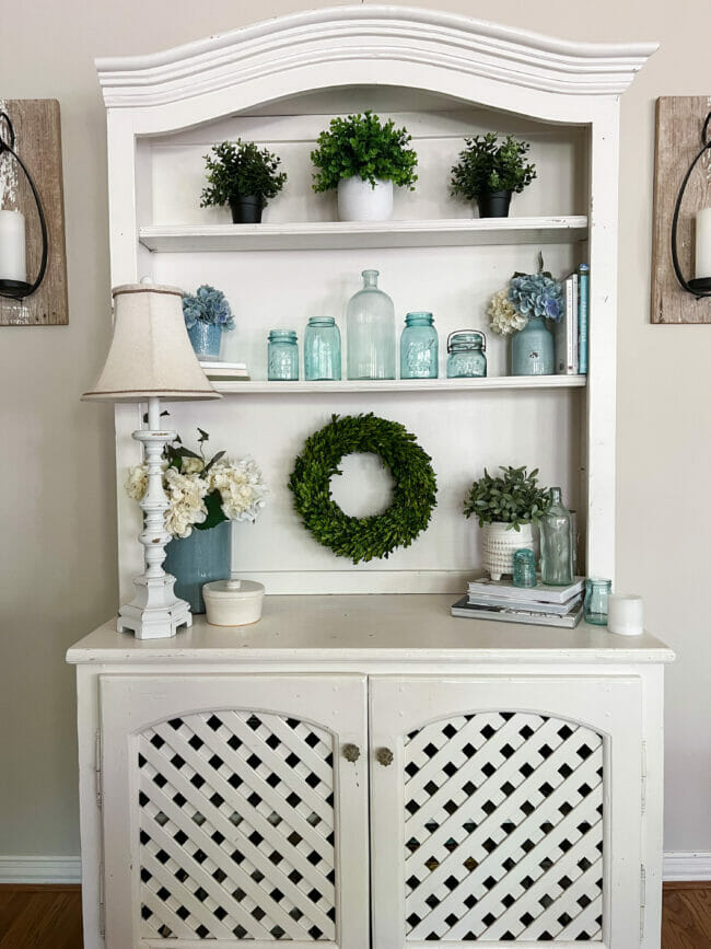 white hutch with wreath, blue bottles, lamp and blue flowers
