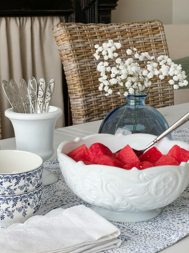 milk glass serving bowl with watermelon, blue vase with white babys breath and utensils and small blue and white bowls