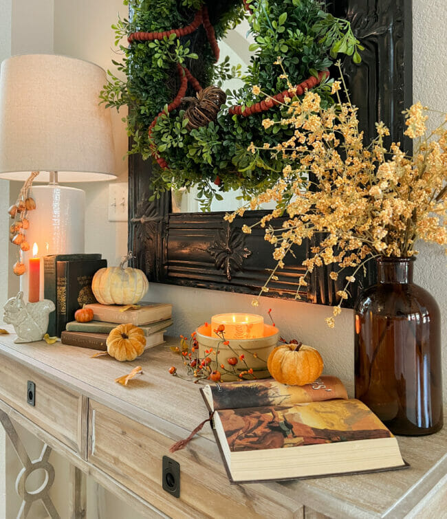 console table with books, pumpkins, brown jar with stems and a lamp