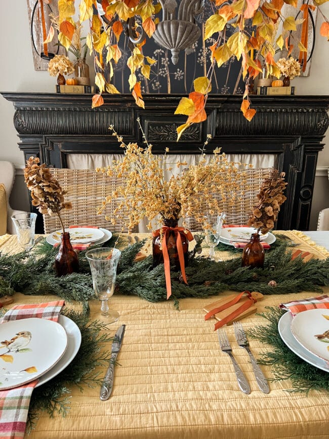 full table with leaves on chandelier, black mantel in background and a brown bottle centerpiece with gold stems