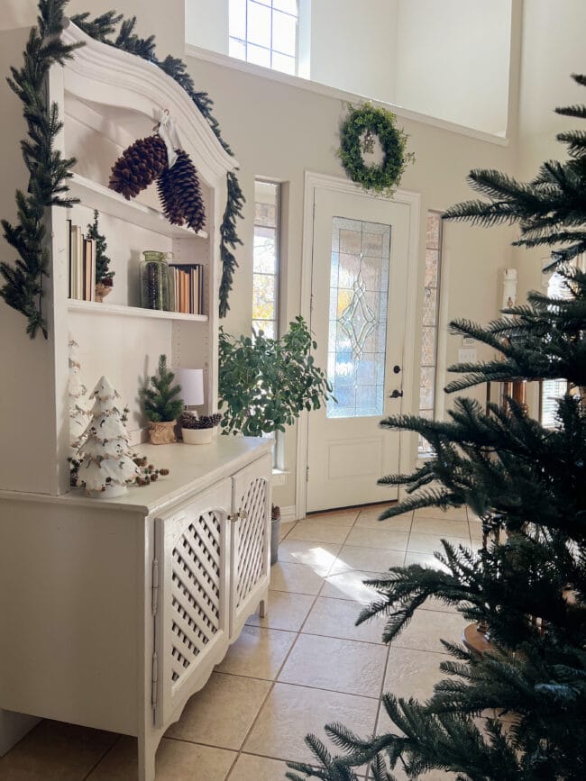 white hutch with Christmas decor, front door with wreath on top and edge of a naked tree