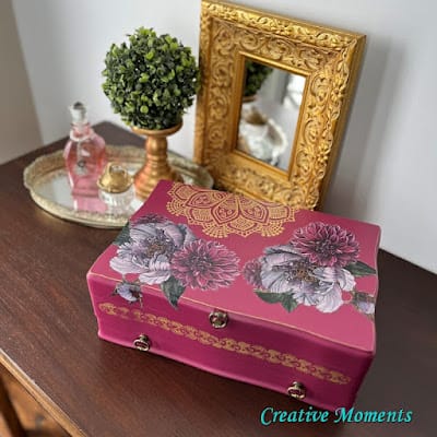 pink crafted jewelry box with gold framed mirror and bottles in background