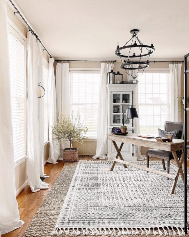 layered rugs with wood desk, white curtains and a plant in corner