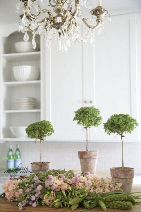 3 topiaries on table with flowers and kitchen cabinets in background