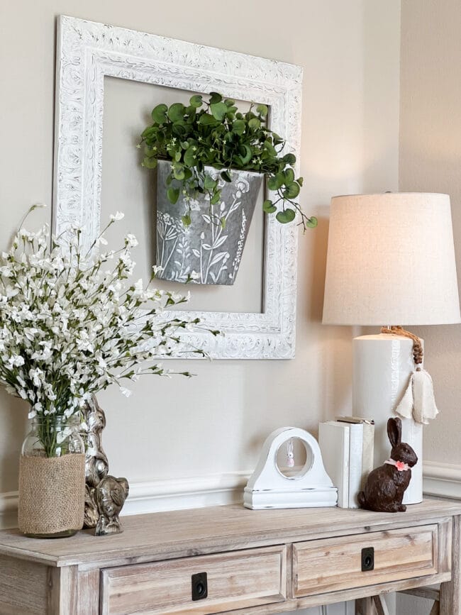 console table with lamp, chocolate bunny and hanging metal basket with greenery inside frame on wall