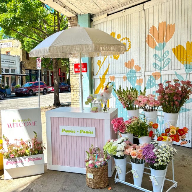 pink cart with umbrella and a flower cart with buckets of pink, orange, purple and white flowers and a welcome sign with flowers. On the street with cars and a tree