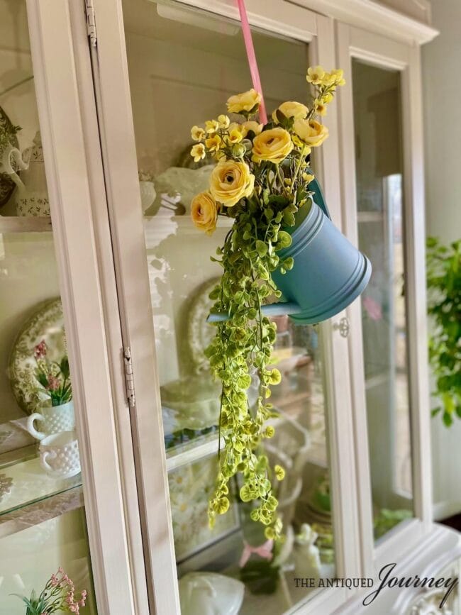blue watering can with yellow flowers and greenery hanging on a dining cabinet
