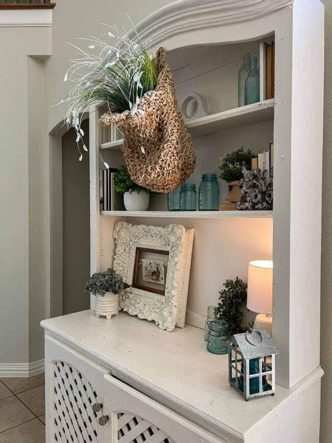 white hutch with blue jars, hanging basket of flowers, books and a large ornate frame