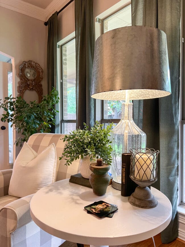 white table in front of windows with glass lamp, faux plant, candle and a vintage green book and table leg