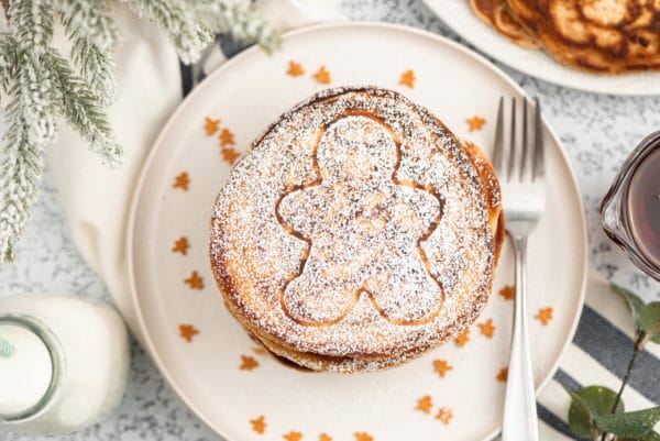 stack of round pancakes with a gingerbread man shape on top on a white plate sprinkled with teeny gingerbread and powdered sugar