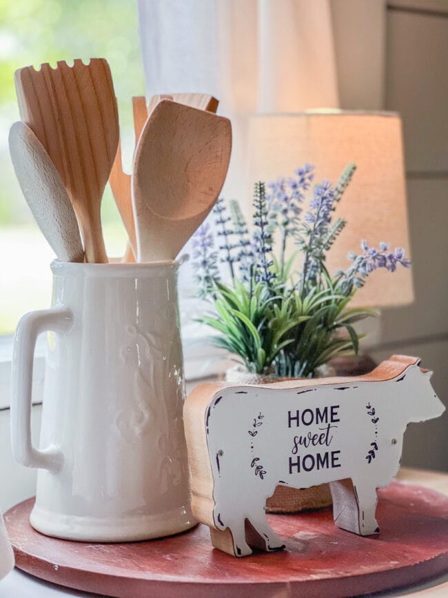 white vintage pitcher with wooden utensils grouped with a bluebonnet plant and cow figurine that says Home sweet home.