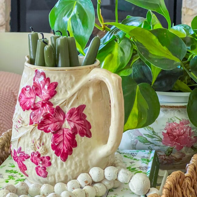 close up of vintage pitcher with pink leaves and green candles inside and plant and beads in background