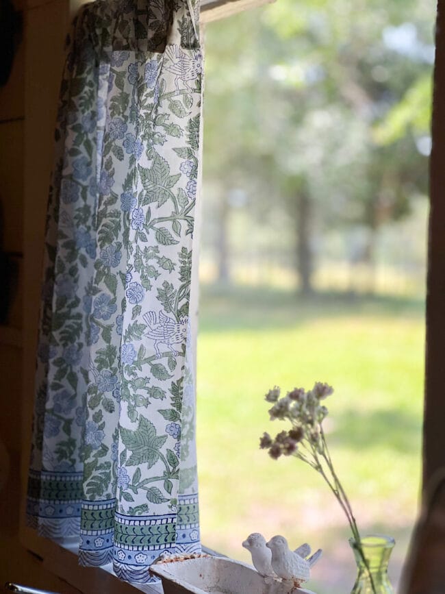 Blue and green curtain pushed open in a window with a small green vase of wildflowers.