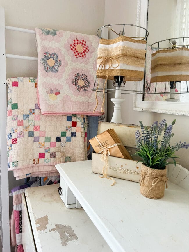 Vintage quilts hanging on ladder with handmade lamp, mailbox with vintage books and a faux lavender plant sitting on a chippy white table