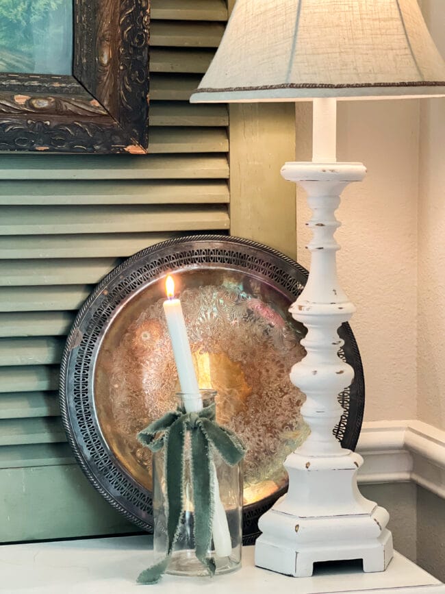 vignette with a silver platter, white lamp and a candle sitting in a clear jar with a green bow tied around the jar.