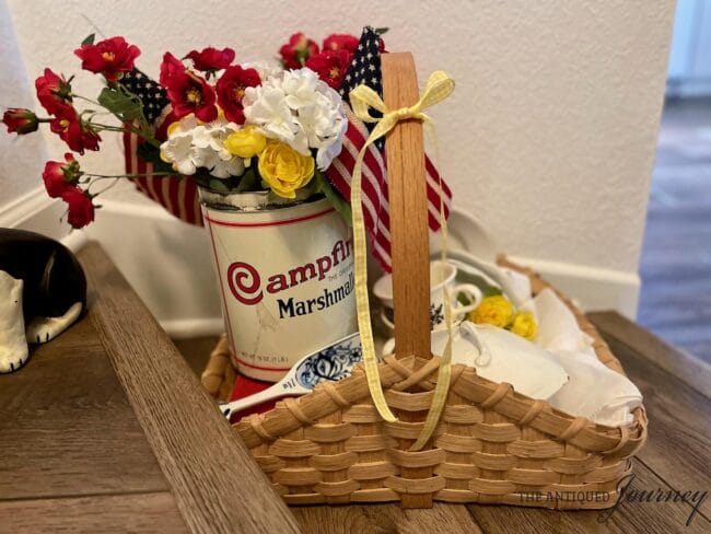 basket with campfire can holding red, white and yellow flowers, a book, vintage dishes, linens, and a blue and white scoop