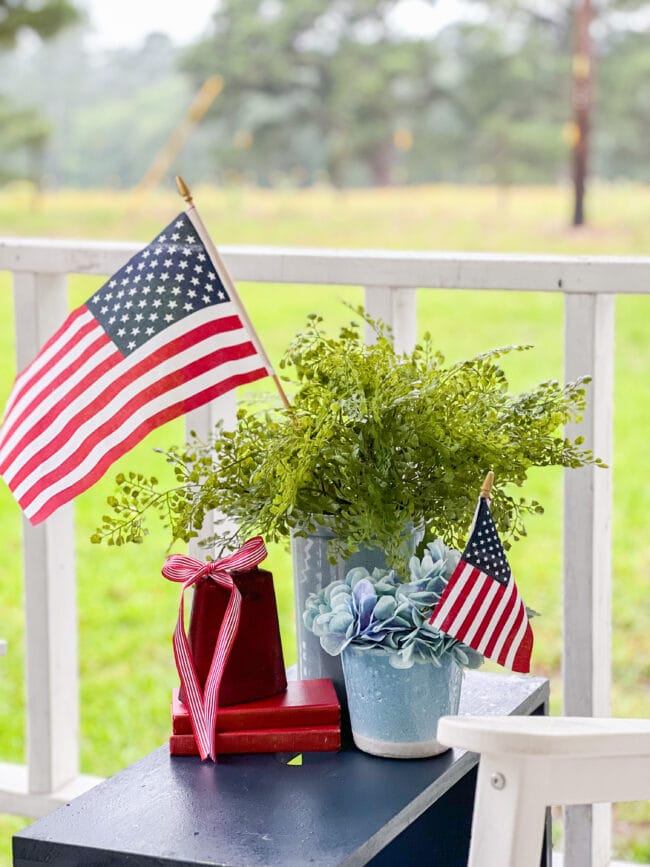 vintage stool with a red bell, books, flags and faux greenery sitting on a porch
