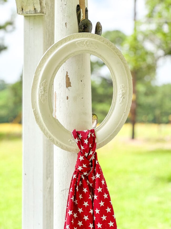 white column sitting outdoors on a porch with a vintage empty frame. A red starry scarf is tied around the frame.