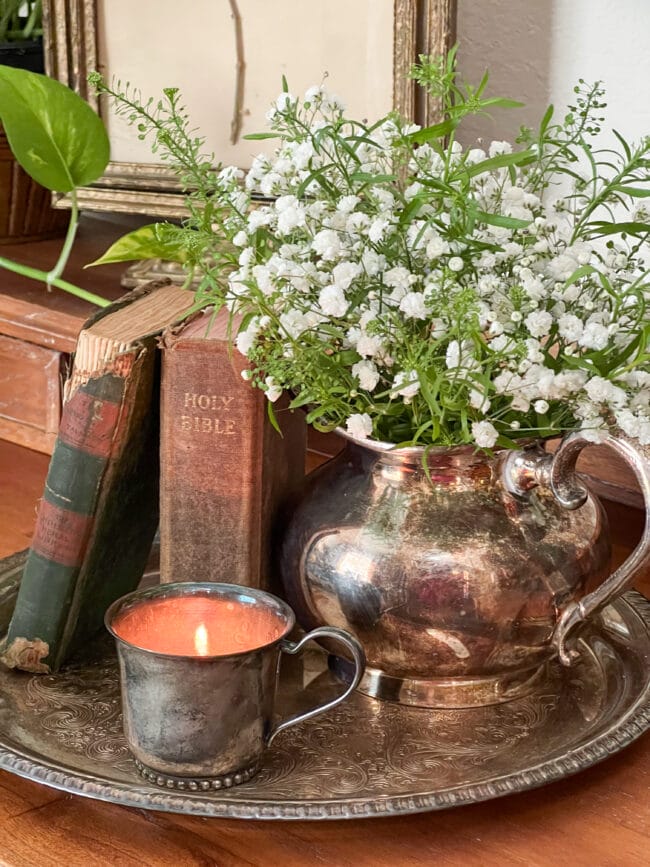 vignette with a silver platter, silver pot with flowers, a baby cup with a candle and vintage books.