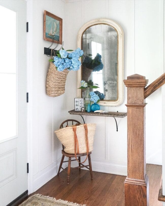 white walls with a stair banister and the corner decorated with a wall mirror, a small chair with a handbag sitting in it and hooks on the wall with a basket of flowers