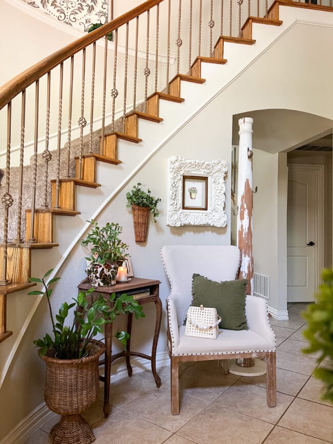 curved staircase with an entryway filled with a chair, column with hooks, side table and plants.