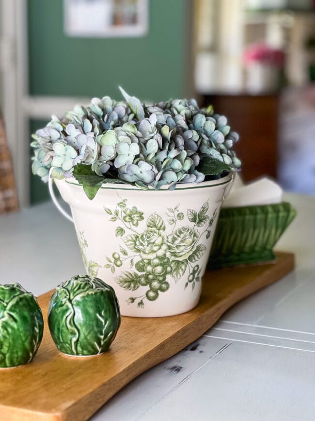 green cabbage salt and pepper shakers, green and white ice bucket with blue hydrangeas and a green dish with napkins all sitting on a cutting board in the center of a table