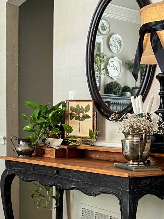 black and wood table with an oval mirror, plants, frames and candles sitting on top