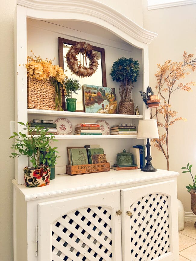 white hutch styled for fall with books, greenery, plates, lamp and an autumn landscape painting.