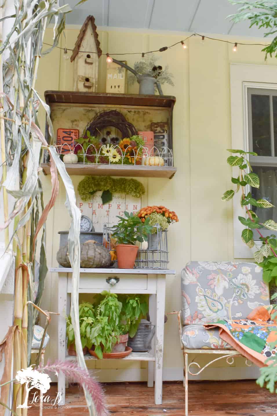 porch with farmhouse table stacked with pots of fall florals, a crate hanging above it with gardening tools, 