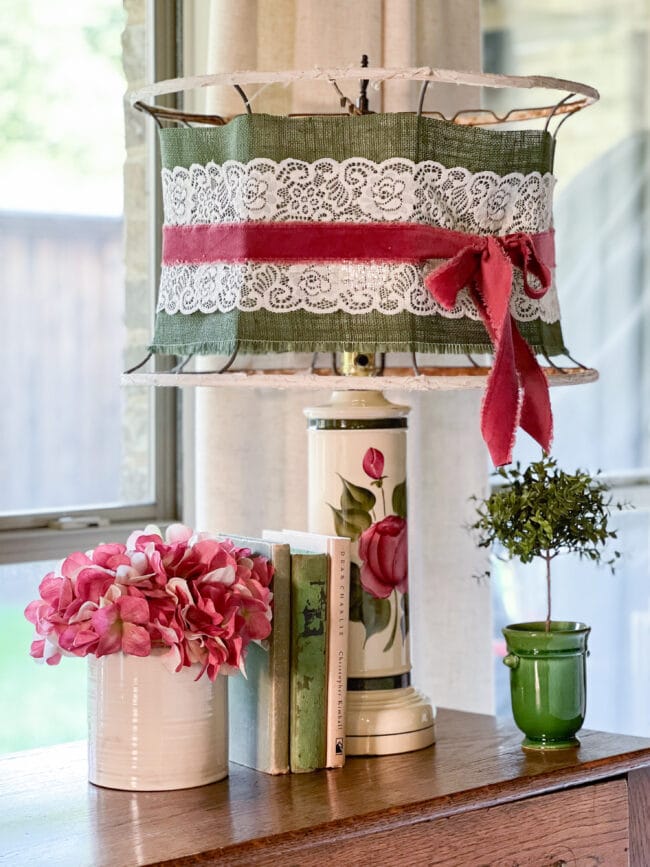 pink and green lamp with floral base and a hand made oversized shade with ribbon. Books, pink flowers and a small green topiary round out the vignette