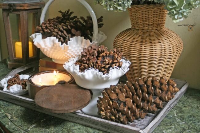 wooden tray full of white milkglass and pinecones with a lit candle.