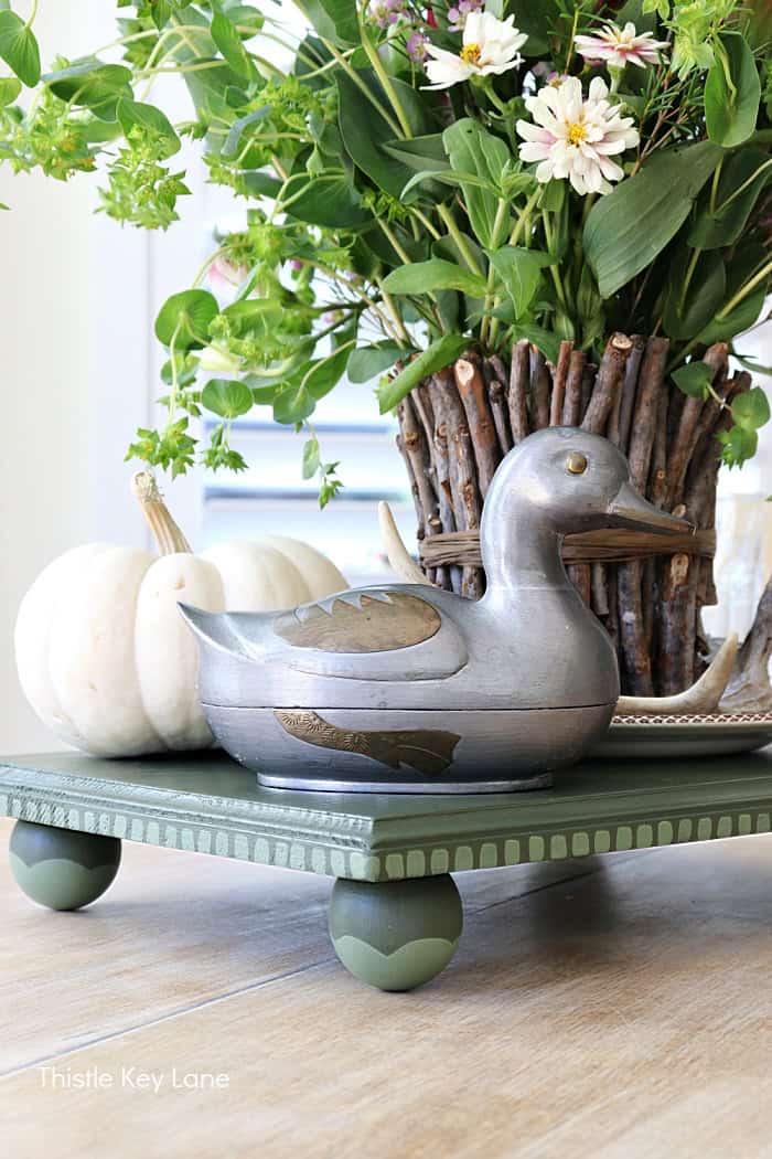 silver and brass duck with pumpkin and plant sitting on a handmade green wooden riser