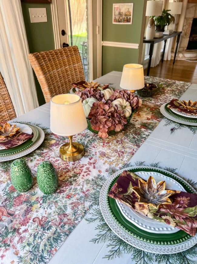 Forest table runner with woodland animals, two tabletop lamps, green salt and pepper shakers, a floral and pumpkin centerpiece, and coordinating place settings.
