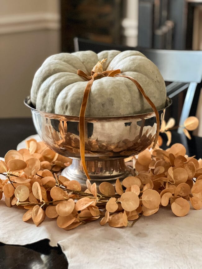 green pumpkin sitting in a footed silver bowl with a gold eucalyptus wreath around the bottom