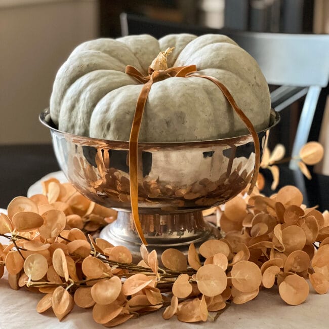 green pumpkin sitting in a silver footed bowl with a gold eucalyptus wreath around the bottom.
