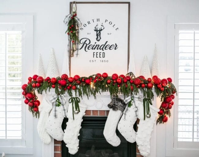 brick mantel with red ornament ball garland, greenery, a reindeer feed print and white stockings.