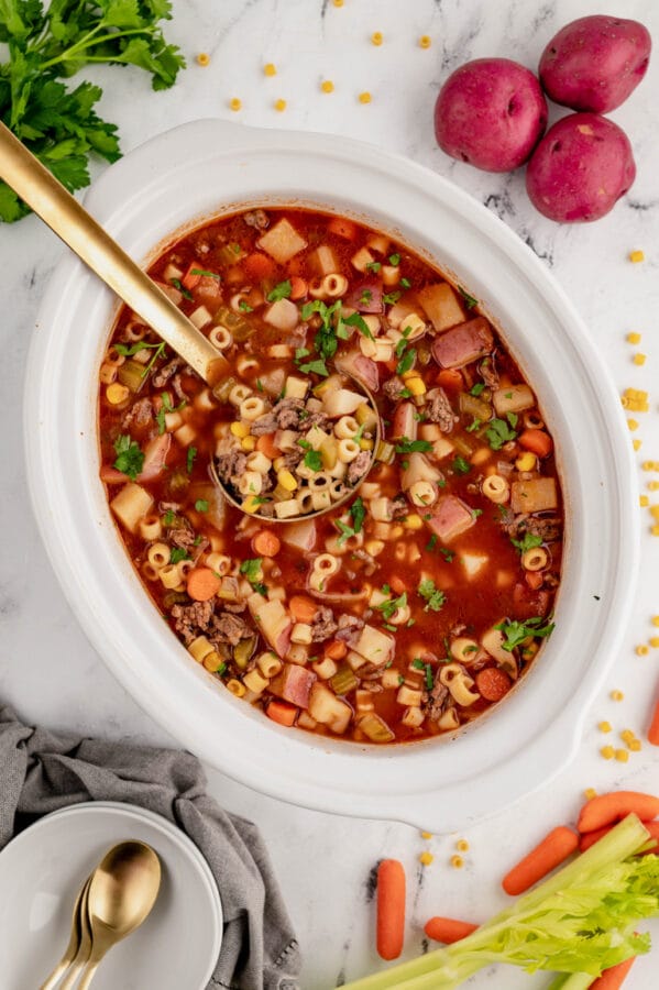 white crock pot of beef and veggie soup with a gold ladle 