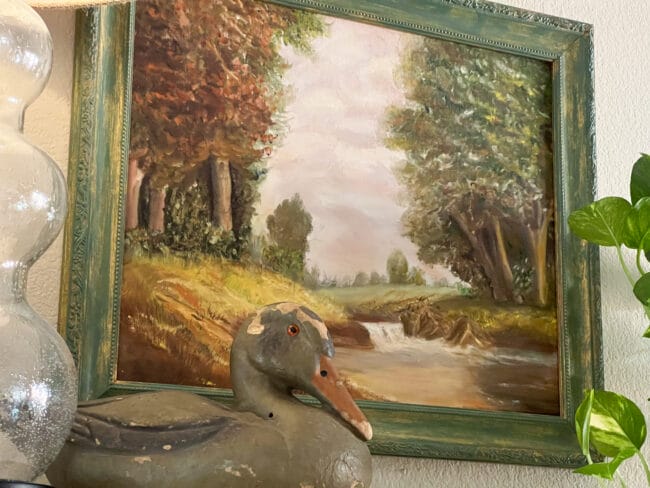 vintage duck sitting in front of a fall colors pastoral painting with trees and a creek